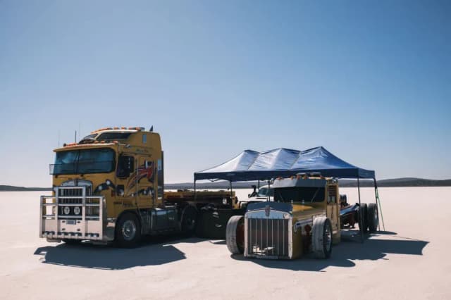 Two vintage yellow Kenworth trucks and a blue pop-up marquee sit parked on the vast white salt lake bed.