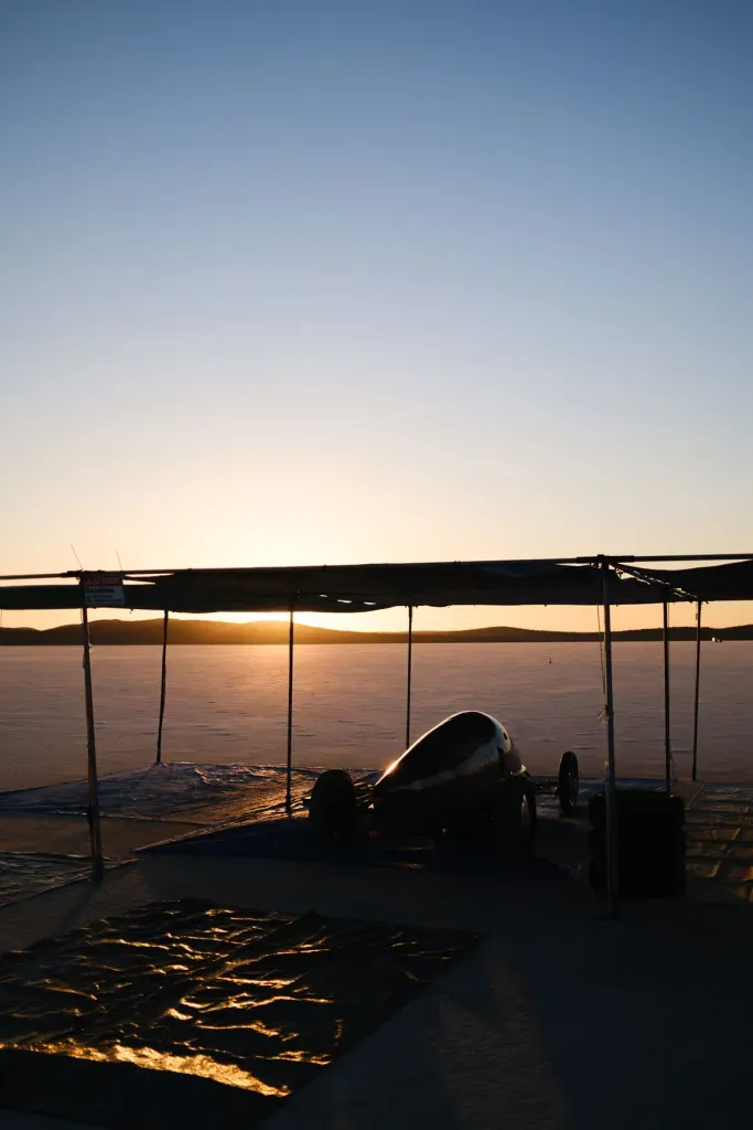 A land speed racing streamliner rests under a canopy on the salt flats as the sun sets.