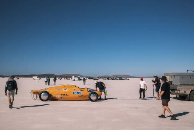 An orange land speed racing car sits on a white salt flat as crew gather around it.