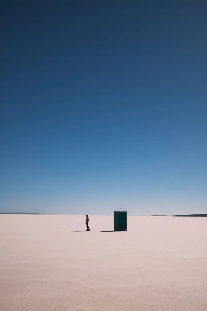 A lone figure stands on a white salt lake beside a portable toilet under a clear blue sky.