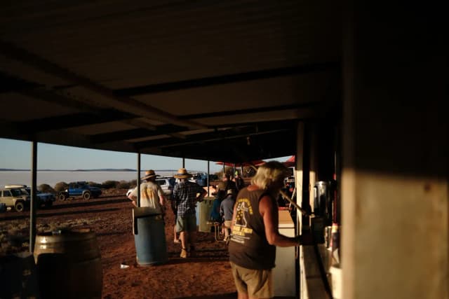 People gather under a tin shed shelter beside a white salt lake at sunset.