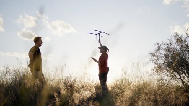 Two men stand in tall grass as one holds up a wildlife tracking antenna against the sky.