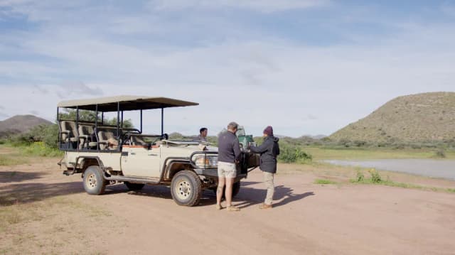A safari guide serves drinks beside an open-top jeep on the savanna, with hills and a river beyond.