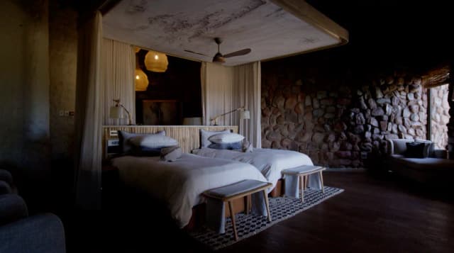 A safari lodge bedroom with two beds, sheer curtains, and a stone feature wall in South Africa.