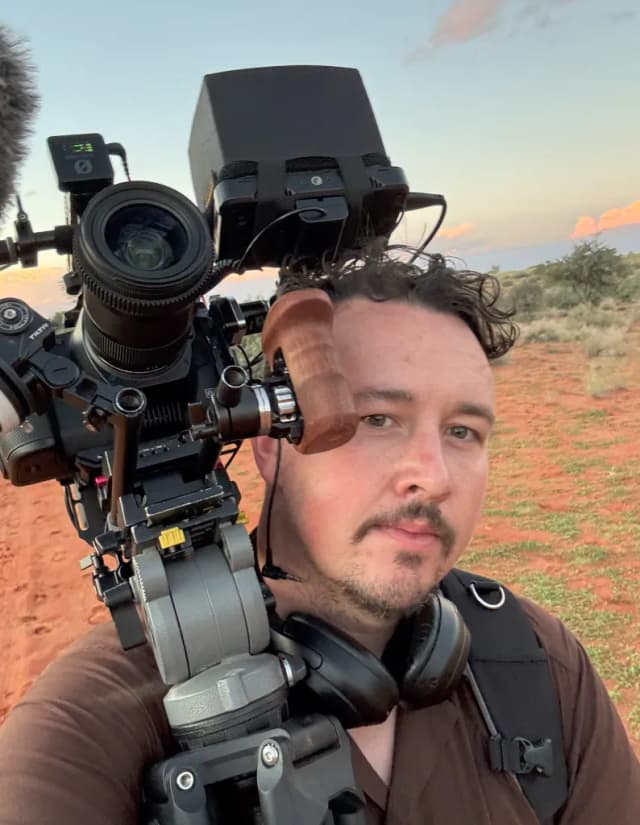 A filmmaker holds a shoulder-mounted camera rig in a red desert landscape at dusk in South Africa.