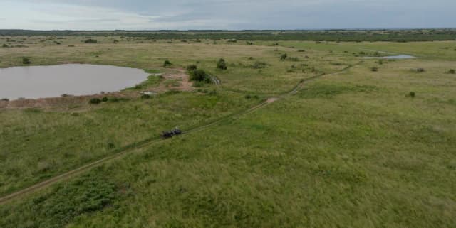 An aerial view of a green wetland landscape with a vehicle driving along a dirt track beside a waterhole.