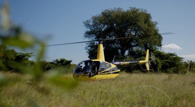 A small yellow Robinson R44 helicopter hovers low over grassland with trees in the background, South Africa.