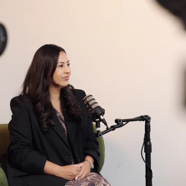 A woman in a black blazer speaks into a boom microphone during a podcast style interview on a green couch.