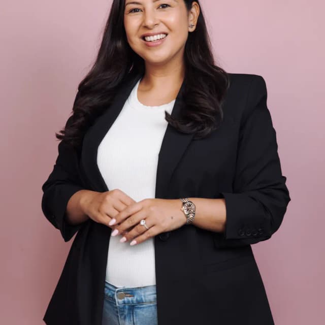 A smiling woman in a black blazer poses against a pink background with her hands clasped.