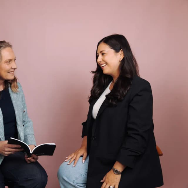 Two women sit and laugh together against a pink backdrop, one holding an open notebook.