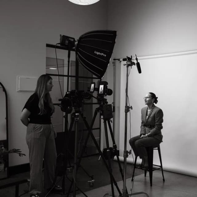 A woman in a black blazer speaks to camera in a bright studio beside a fiddle leaf fig plant.
