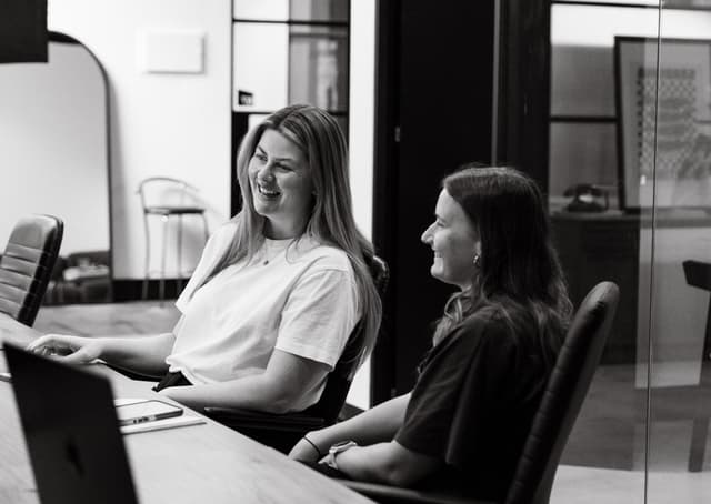 Two women laugh together while looking at a laptop screen in a black and white office photo.