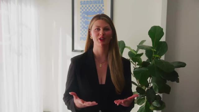 A woman in a black outfit speaks to camera in a bright office beside a leafy plant, an advertising campaign visual.