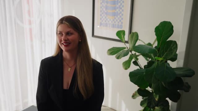 A smiling woman in a black blazer stands near a potted fig plant and a framed motorsport poster.