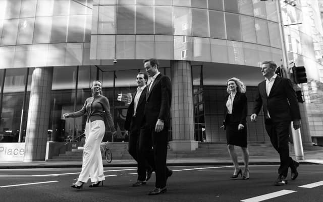 A black and white photo captures five colleagues in business attire crossing a city street outside a glass office building.
