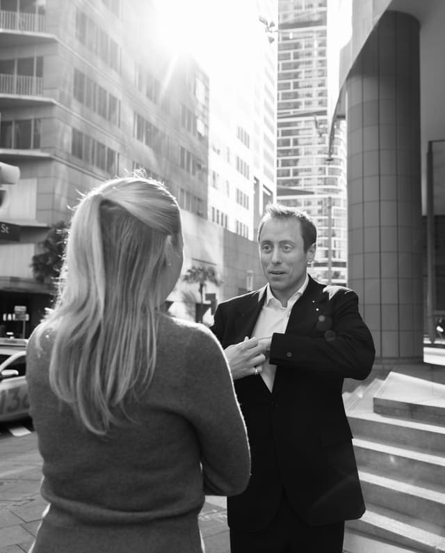 In black and white, a man and woman speak together on a sunlit city street lined with high-rises.