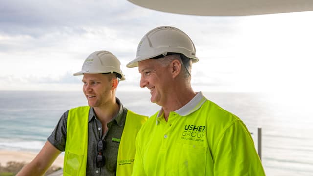 Two Usher Group employees in hard hats and hi-vis vests smile together with the ocean behind them.