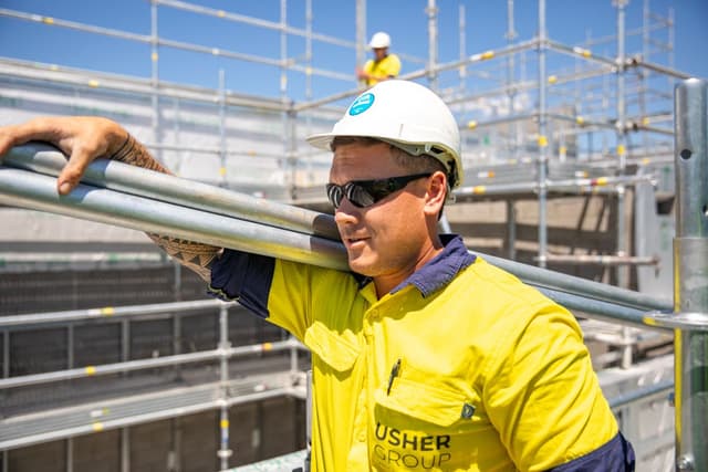 A tradesperson in a yellow Usher Group hi-vis shirt holds scaffolding poles on a construction site, wearing a hard hat and sunglasses.