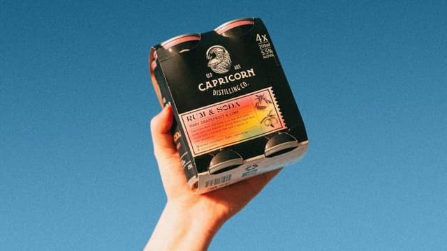 A hand holds up a four-pack of Capricorn Distilling Co rum and soda cans against a blue sky.