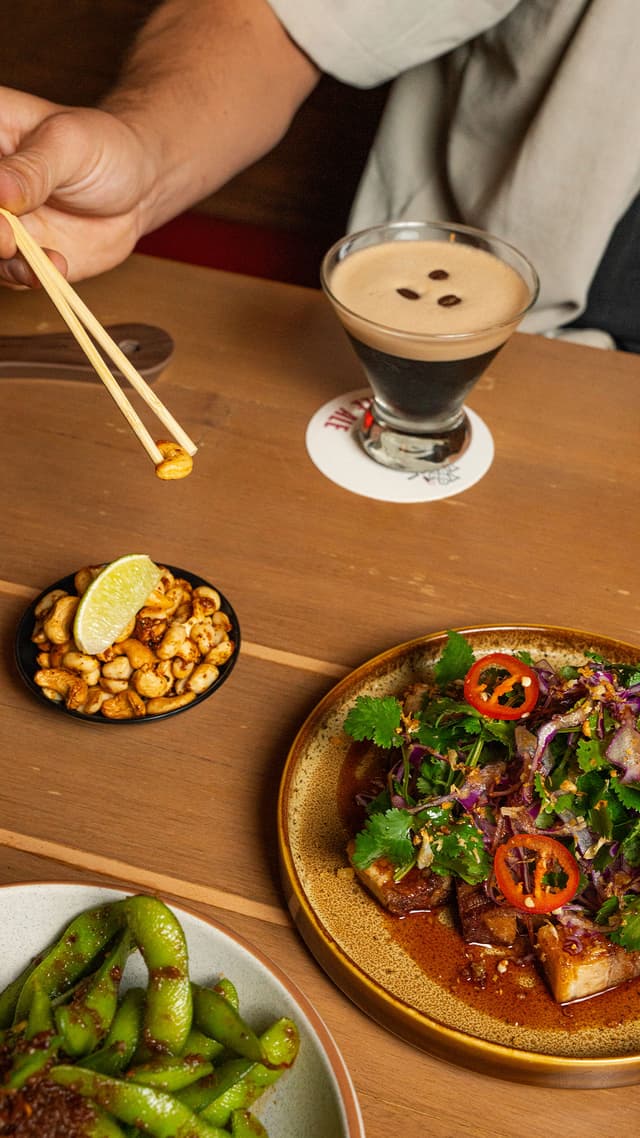 A diner picks up a spiced cashew with chopsticks beside an espresso martini, pork belly, and edamame on a restaurant table.