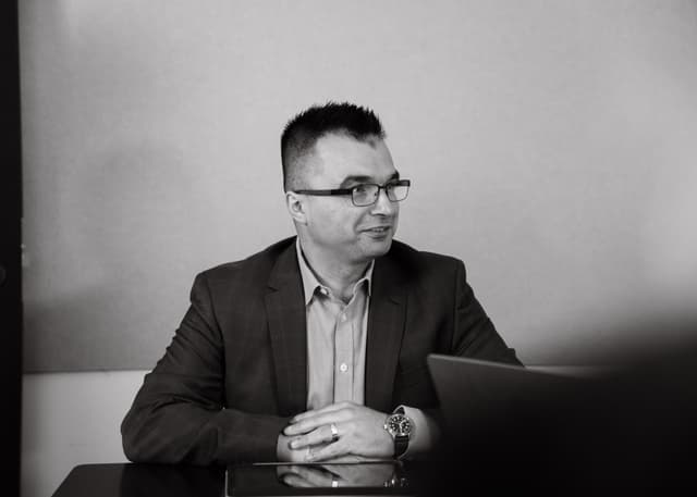 Black and white portrait of a man in a blazer and glasses sitting at a desk with a laptop.