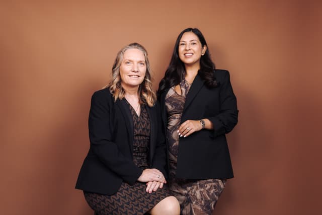 Two women in dark blazers pose together for a studio portrait against a warm brown backdrop.