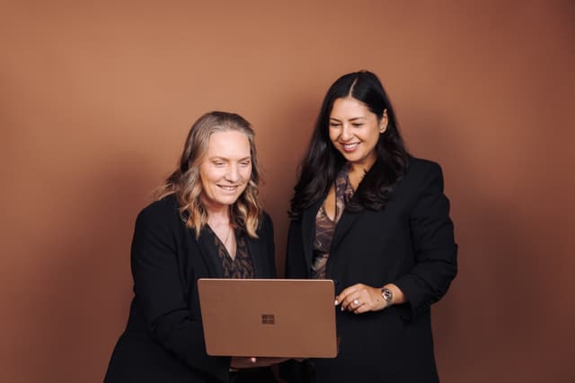 Two women smile while looking together at a laptop screen against a warm brown backdrop.