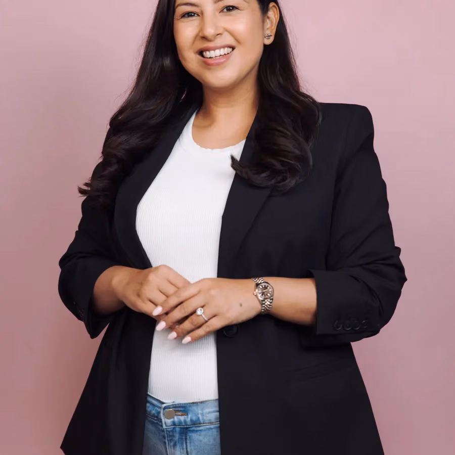 A smiling woman in a black blazer and white top poses for a portrait against a soft pink backdrop.