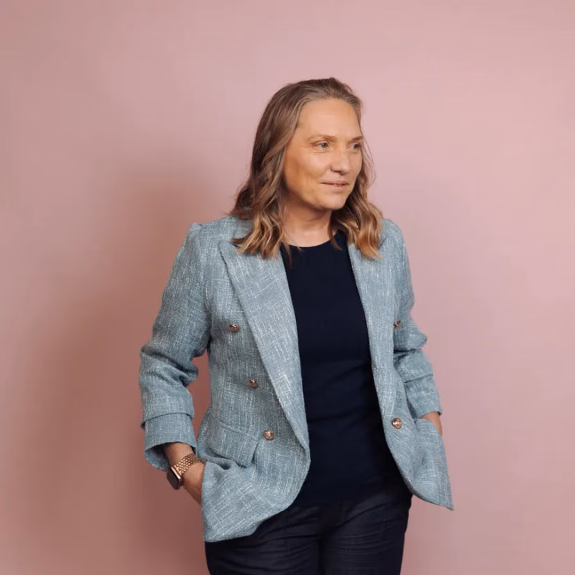 A woman in a sage green blazer poses for a studio portrait against a dusty pink backdrop.