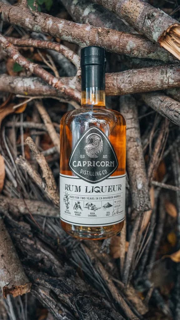 A bottle of Capricorn Distilling Co rum liqueur rests among dry branches, its label reading rum liqueur aged in ex-bourbon barrels.