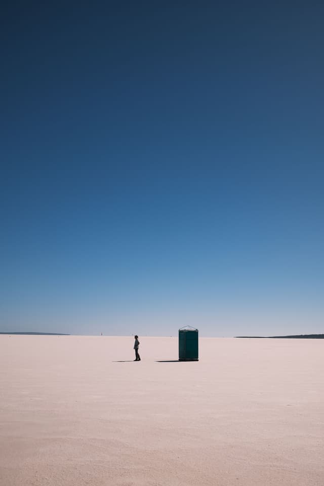 A lone figure stands on a vast white salt lake beside a portable toilet under a deep blue sky.