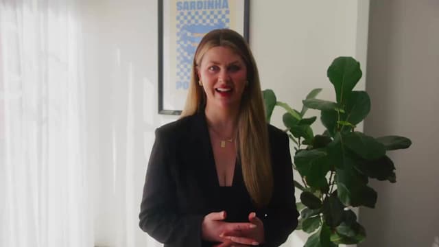 A woman in a black outfit speaks to camera in a bright office beside a leafy plant and framed poster.