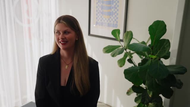 A woman smiles while speaking to camera in a bright office beside a leafy plant.