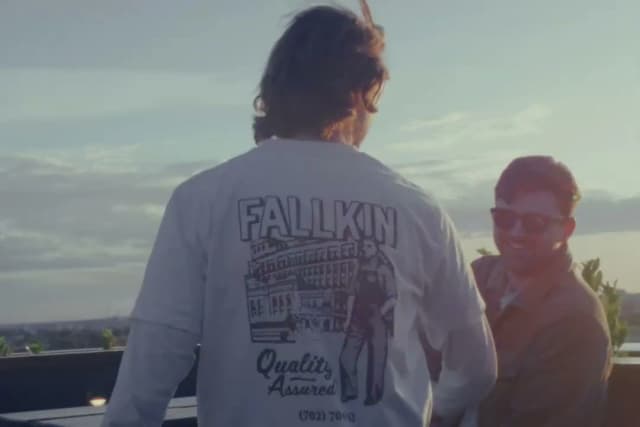 Two men on a rooftop at sunset, one wearing a Fallkin graphic shirt, laughing together.