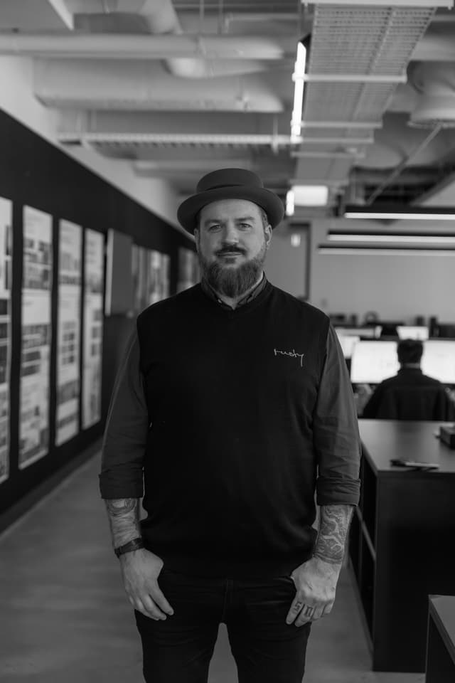 A black and white portrait of a bearded man in a hat and dark sweater in an office corridor with project boards.
