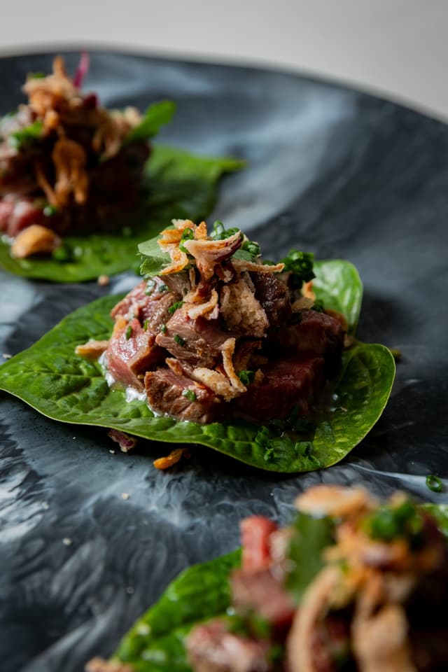 Bite-sized beef pieces topped with crispy shallots and chives, served on individual leaves on a dark plate.
