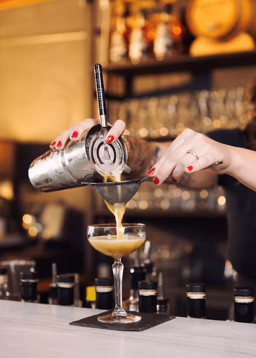 A bartender's hands strain a creamy cocktail into a coupe glass at a softly lit bar.