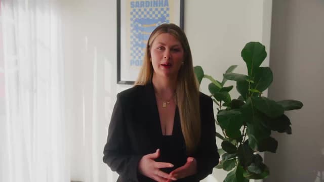 A woman in a black blazer speaks to camera in a bright studio with a fiddle leaf fig and framed poster.