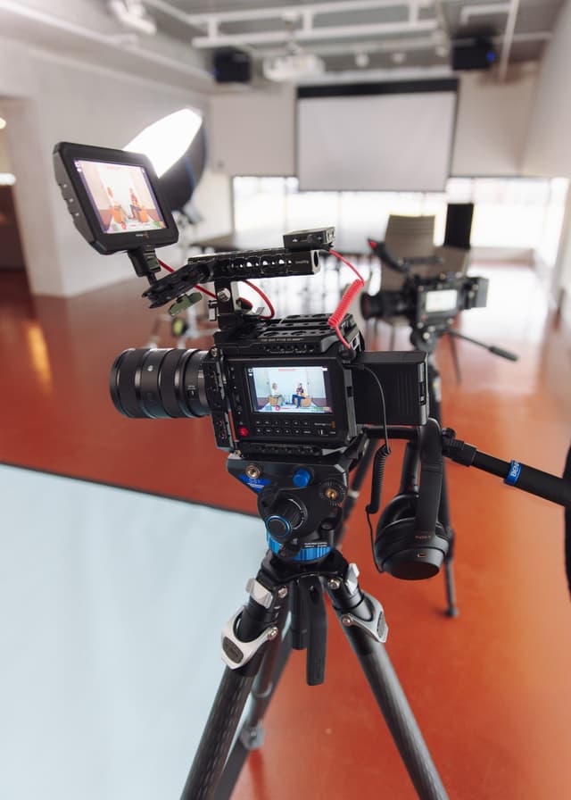 A cinema camera rig with a monitor screen captures two people on a podcast set behind the scenes.