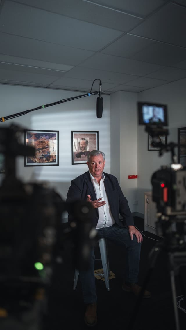 A man is interviewed on a stool in a studio, framed by cameras, a boom microphone, and wall art.