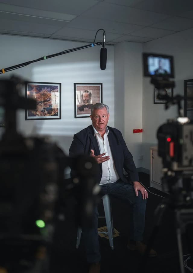 A man sits on a stool answering questions during an on-camera interview, framed between two cinema cameras.