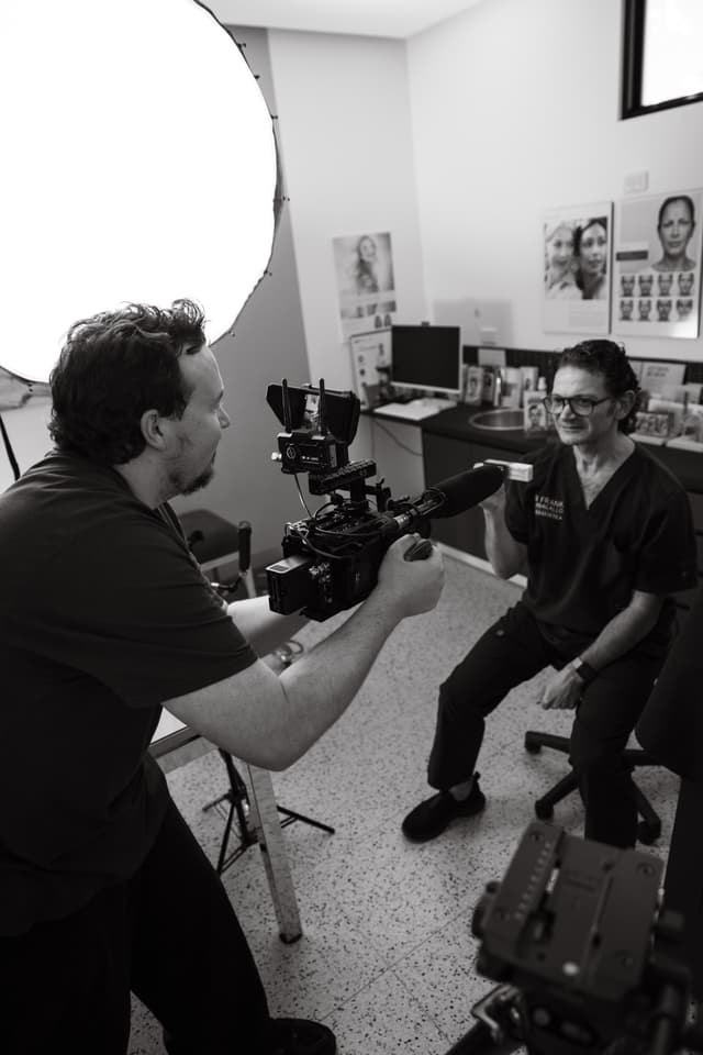 A videographer films a smiling doctor in scrubs seated in a clinic room, a black and white behind the scenes shot.