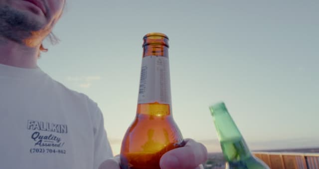 A man wearing a Fallkin branded shirt holds up two beer bottles against a sunset sky on a rooftop.