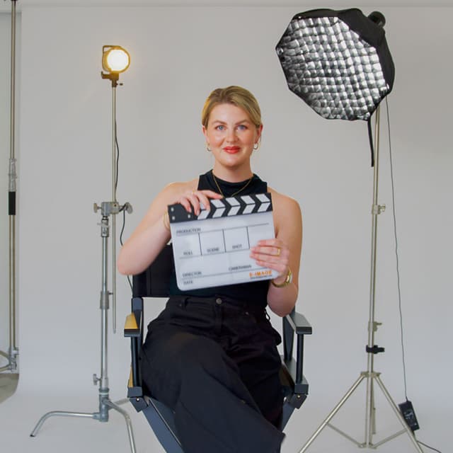 A woman sits in a director's chair holding a clapperboard, framed by studio lights and a softbox.