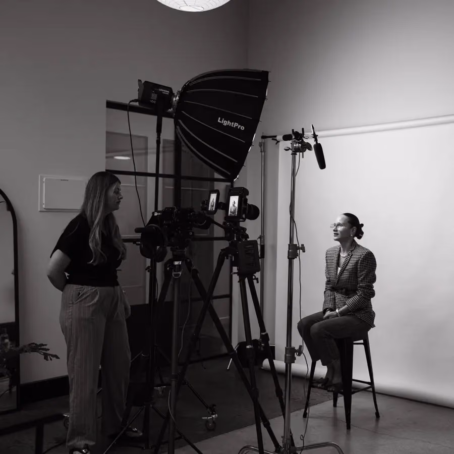 A black and white behind the scenes shot shows a camera crew filming a seated woman under a softbox light.