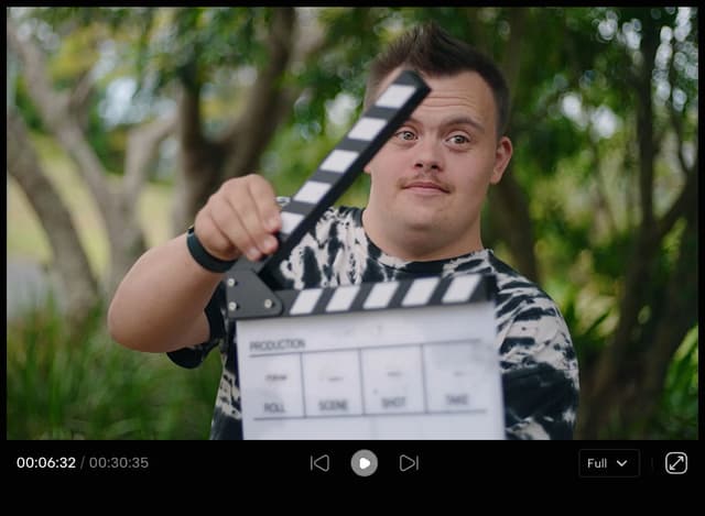 A young man with a clapperboard smiles outdoors in a video player still, part of a video production process.