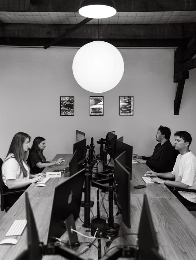 Five colleagues work at a row of computer monitors in a studio lined with framed prints.
