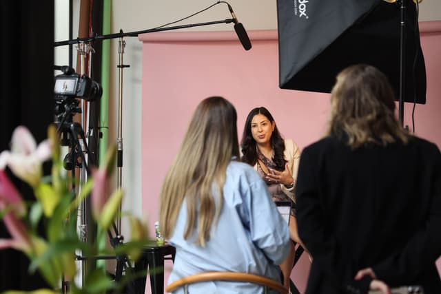 A film crew records a woman being interviewed against a pink backdrop with softbox lighting and a boom microphone.