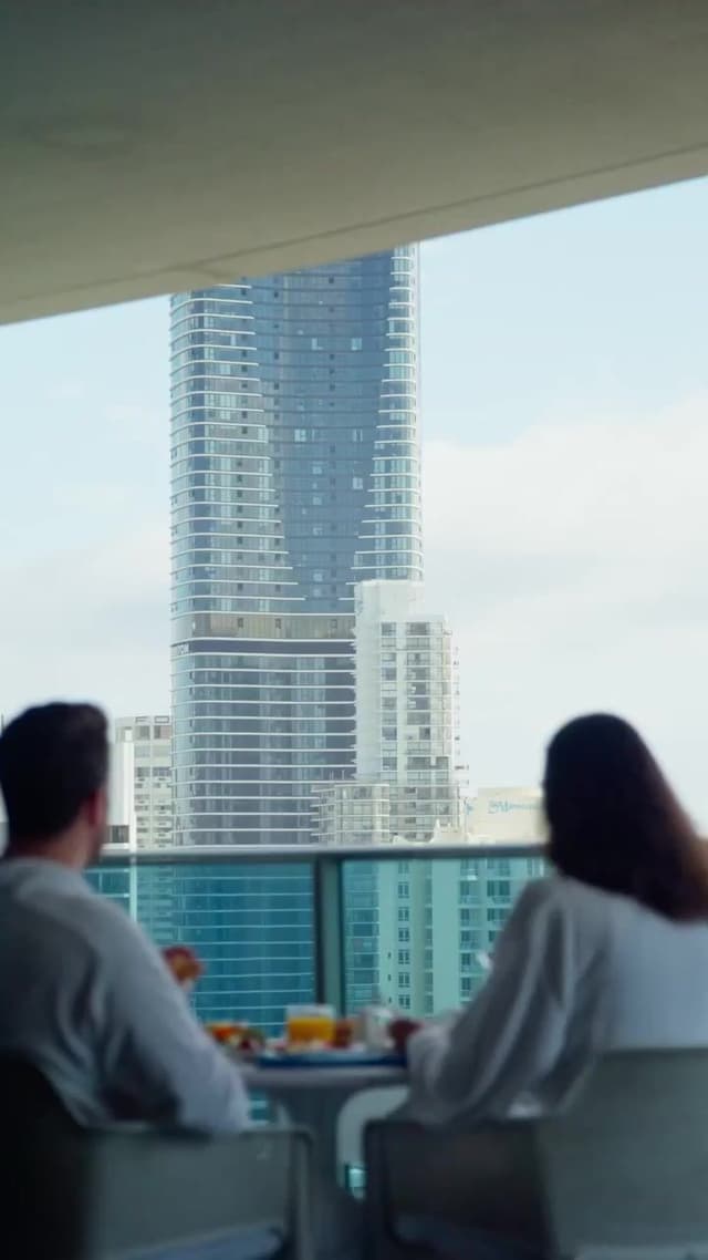 A couple share breakfast on a high rise balcony overlooking coastal towers and the ocean in the distance.