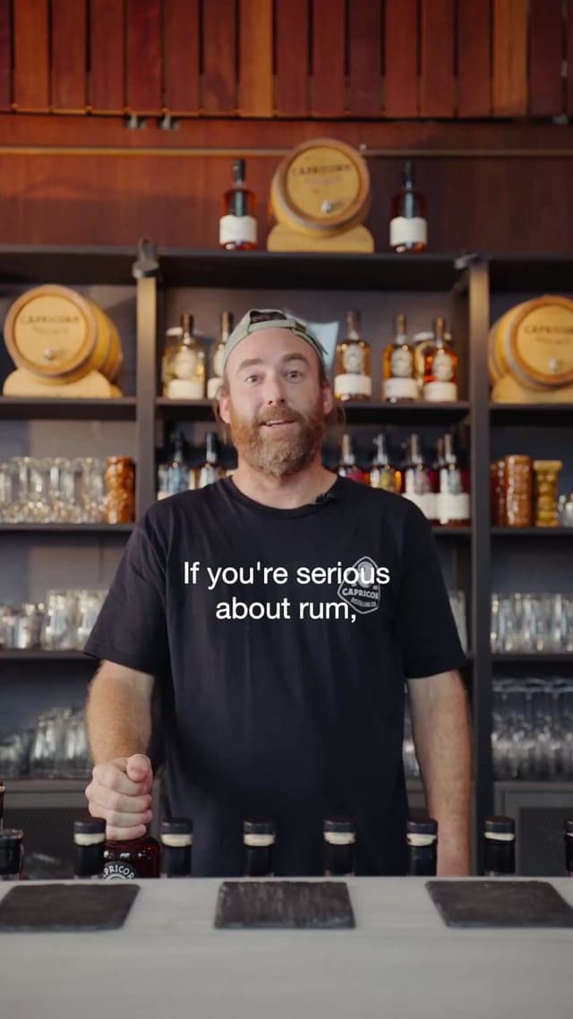 A bearded bartender in a Capricorn branded shirt talks to camera in front of shelves of spirits.